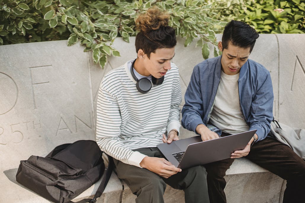 Serious young multiethnic men students in casual clothes studying on laptop while sitting on stone bench in street in daytime with backpack near green plants