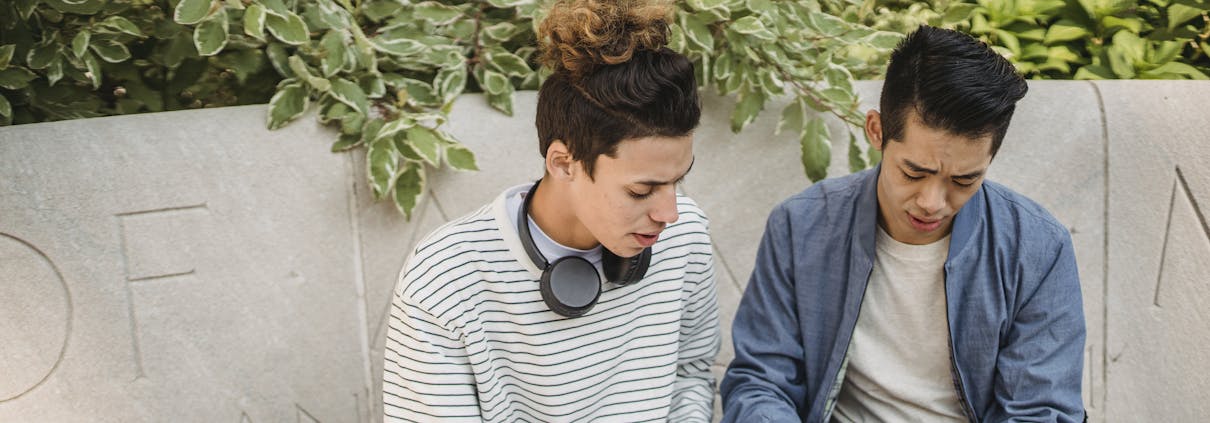 Serious young multiethnic men students in casual clothes studying on laptop while sitting on stone bench in street in daytime with backpack near green plants