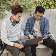 Serious young multiethnic men students in casual clothes studying on laptop while sitting on stone bench in street in daytime with backpack near green plants
