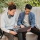 Serious young multiethnic men students in casual clothes studying on laptop while sitting on stone bench in street in daytime with backpack near green plants